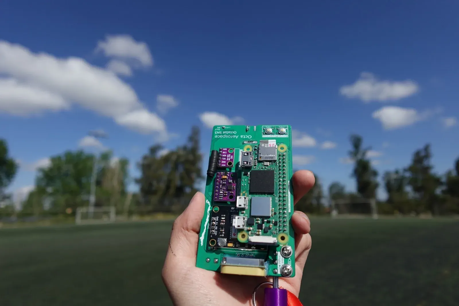 An OctaSat SN3 flight board held up against a blue sky over a football field