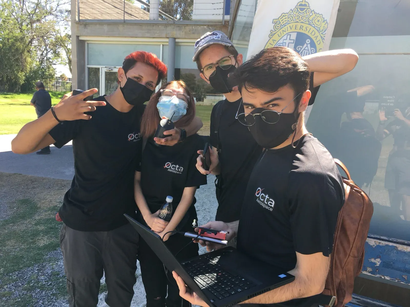 Four team members in Octa Aerospace shirts and face masks on a university campus